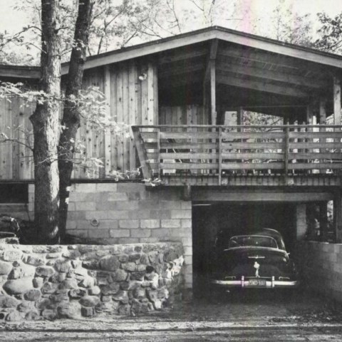 Black and white image of a wooden house on stilts with a vintage car in the garage.