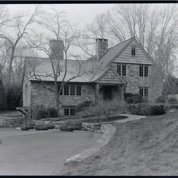 Black and white photo of a stone house with a driveway and bare trees.