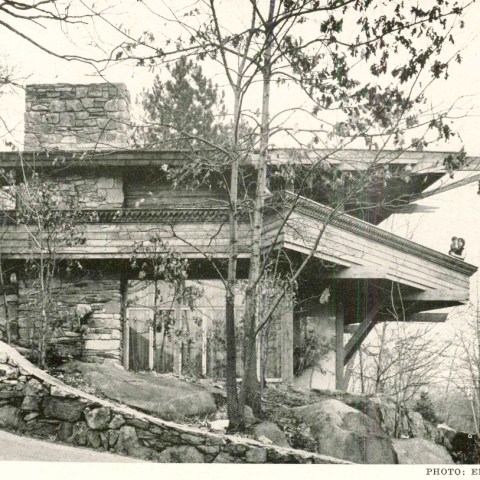 A rustic two-story house with stone and wood, surrounded by trees in a natural setting.