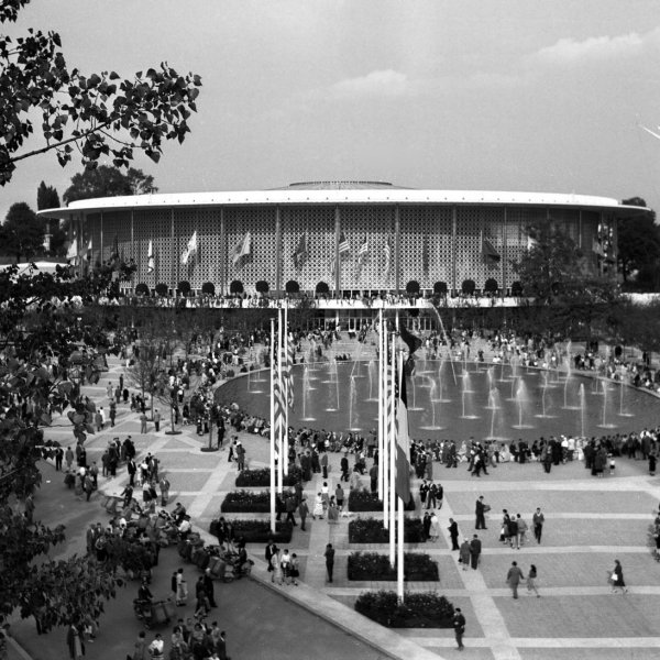 Crowd gathered around a large fountain in front of a round building with flags.