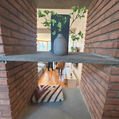 Vase with green leaves on brick shelf, view into a living room with people.