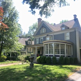 Large gray Victorian house with gabled roof, porch, and trees in sunny yard.