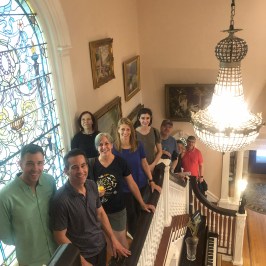 Group of eight people standing on a staircase by stained glass window and chandelier.