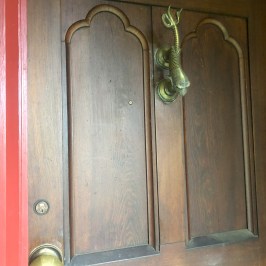 Wooden door with brass door knocker and handle, red frame on the left.