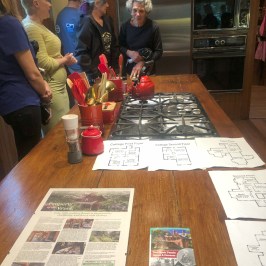 People in a kitchen discussing over maps and pamphlets on a wooden island.