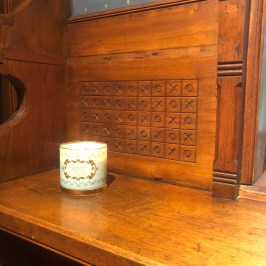 Lit candle on wooden shelf with tic-tac-toe pattern on wall panel behind.