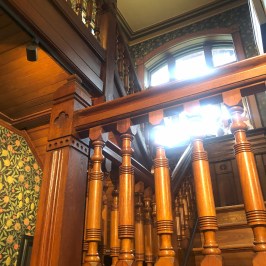 Ornate wooden staircase with floral wallpaper and arched window.