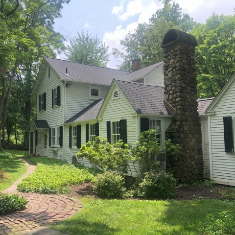 White house with stone chimney, black shutters, and lush greenery surrounding it.