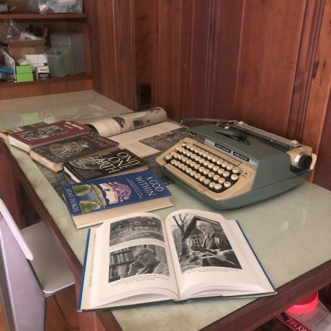 Vintage typewriter on desk with open book, magazines, and wooden wall background.