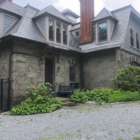 Stone house with gray shingles, red brick chimney, and green plants in the front yard.