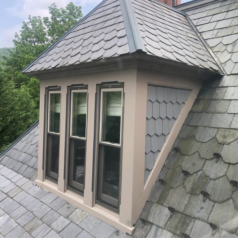 Gabled roof with four vertical windows and slate shingles, red brick chimney in background.