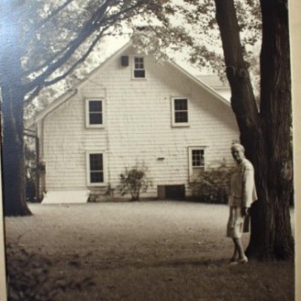 Woman standing by a tree in front of a white wooden house with two windows.