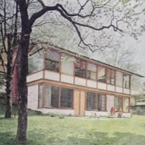 Two-story modern house with large windows, surrounded by trees and grassy lawn.