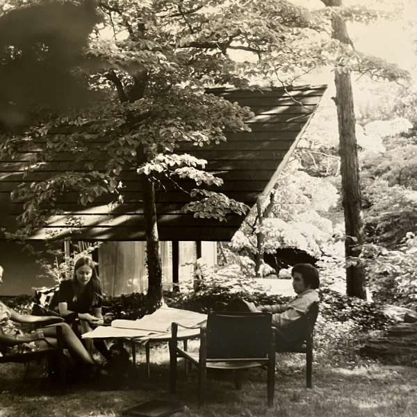 Three people sitting around a table outdoors near a house surrounded by trees.