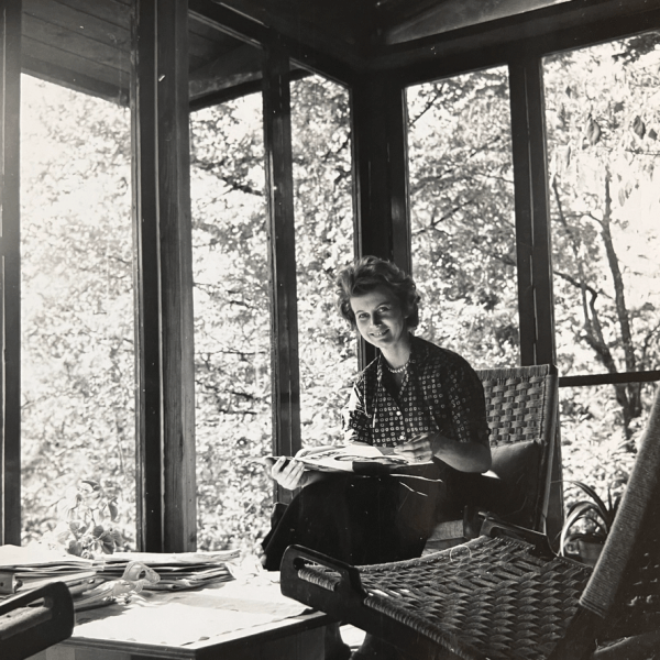 Woman sitting in a sunlit room with large windows, reading a book, surrounded by trees.