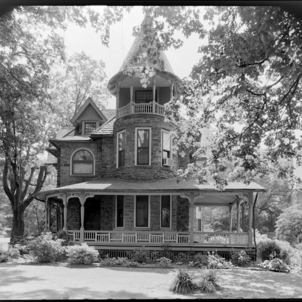Victorian house with turret and wraparound porch, surrounded by trees.