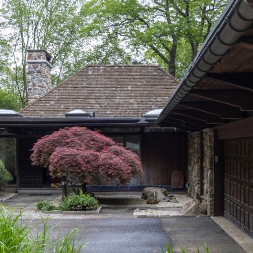 Stone house with chimney, surrounded by lush trees and bushes, featuring a driveway.