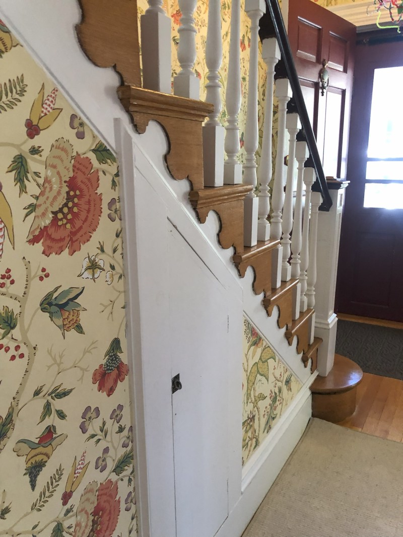 Wooden staircase with floral wallpaper and white spindles near a red door.