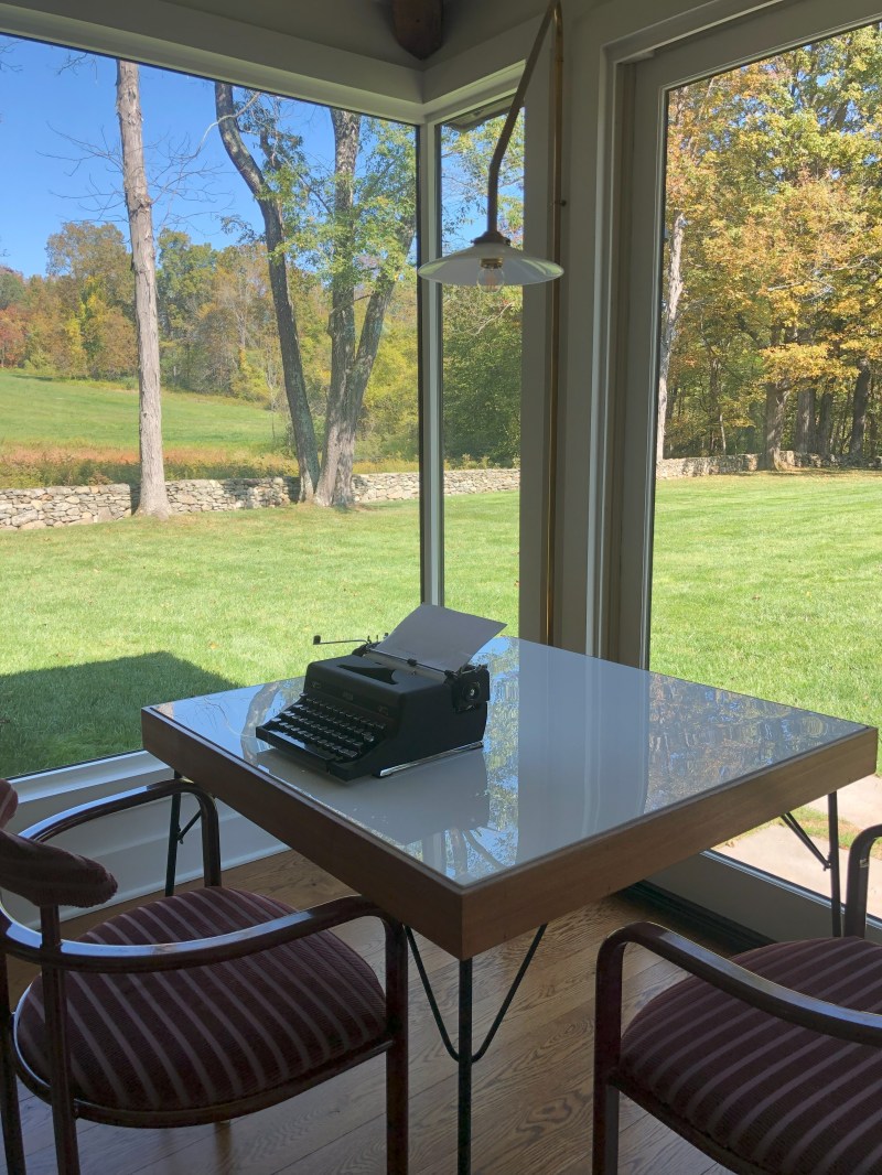 Typewriter on a table by large window overlooking a grassy field and trees.