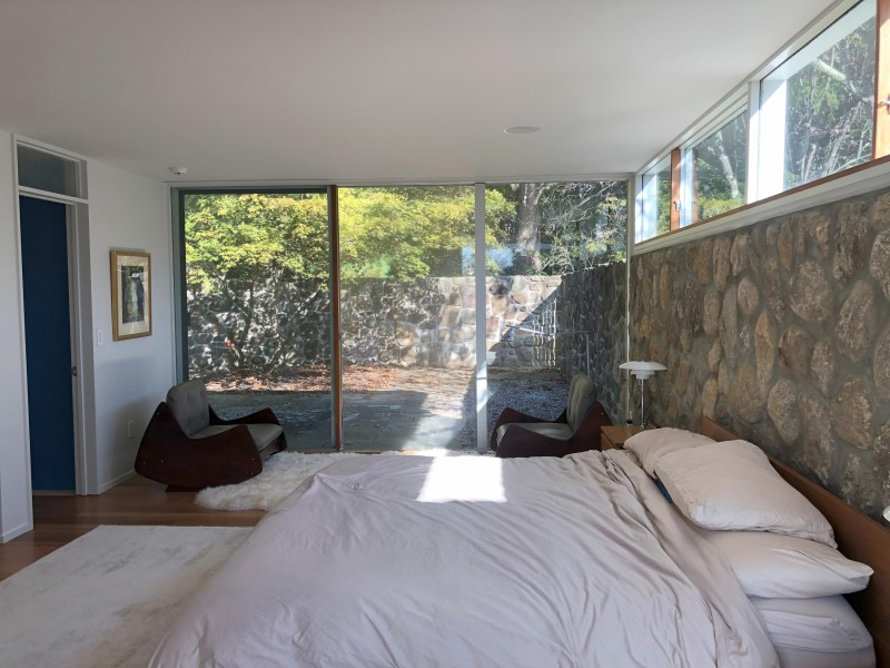 Modern bedroom with large windows, stone wall, and minimalist furniture.
