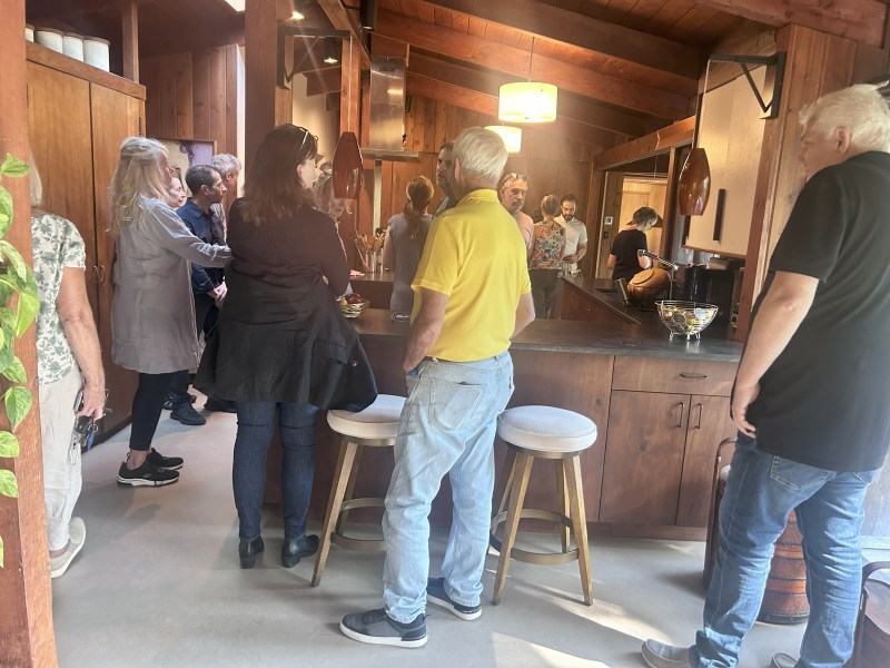 Group of people gathered in a cozy wooden kitchen with stools and pendant lights.