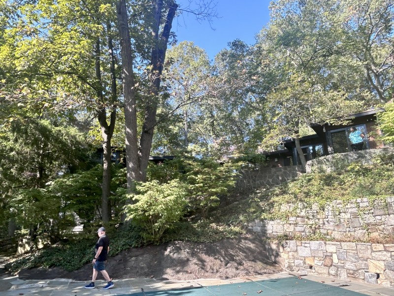 Person walks on a path beside trees and a house on a hill.