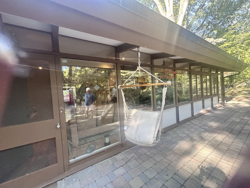 Glassed patio with a hanging chair, surrounded by trees and brick paved floor.
