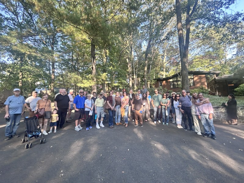 Large group of people posed on a shaded road with trees and a house in the background.