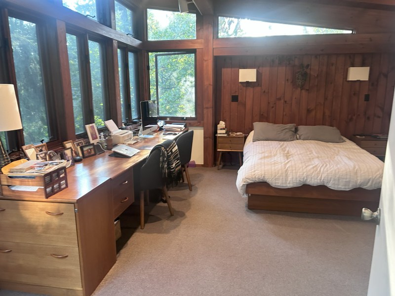 Bedroom with wood walls, a desk by large windows, and a bed with grey pillows.