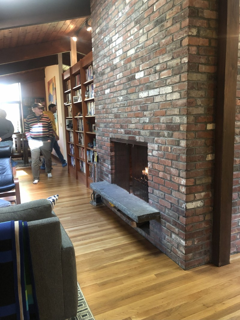 Brick fireplace in cozy room with bookshelves and people walking.