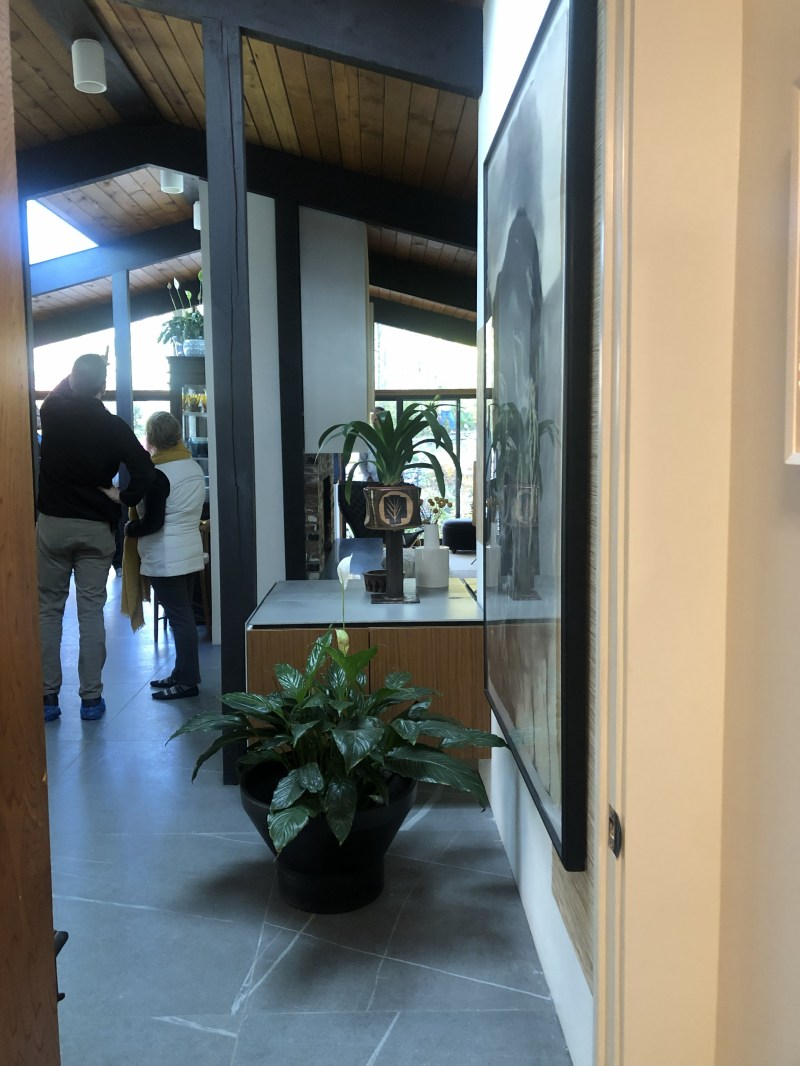 Interior hallway with plants, wooden ceiling, and two people looking out a large window.