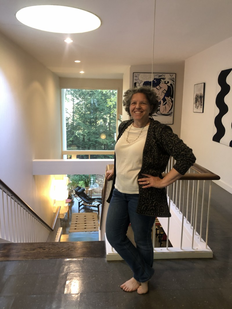 Smiling person standing in a hallway near a staircase, with artwork and large window in the background.