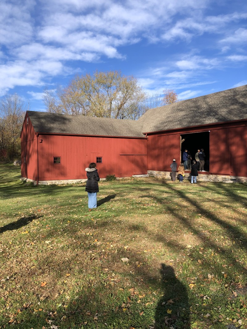 People near a large red barn with a tree-lined background under a partly cloudy sky.
