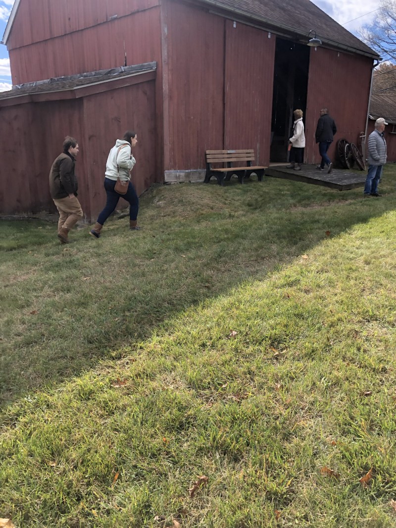 People walking towards a large red barn on a grassy area with a bench nearby.