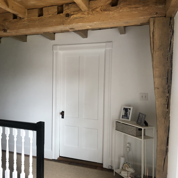 Wood-beamed hallway with a white door and small table beside a staircase.