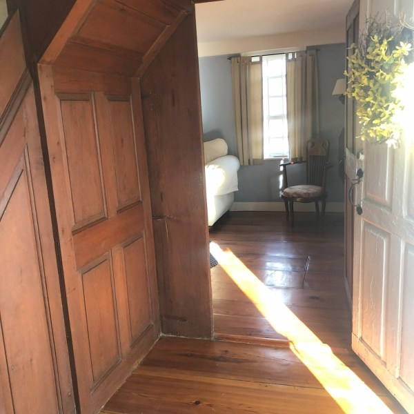 Sunlit wooden hallway leading to a bright room with a bed and chair.